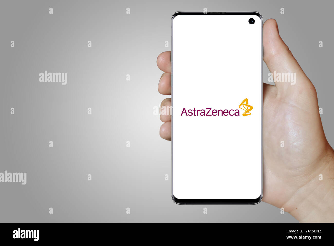 A man holds a smartphone displaying the logo of company AstraZeneca listed on OMX Stockholm. Grey gradient in the background. Credit: PIXDUCE Stock Photo