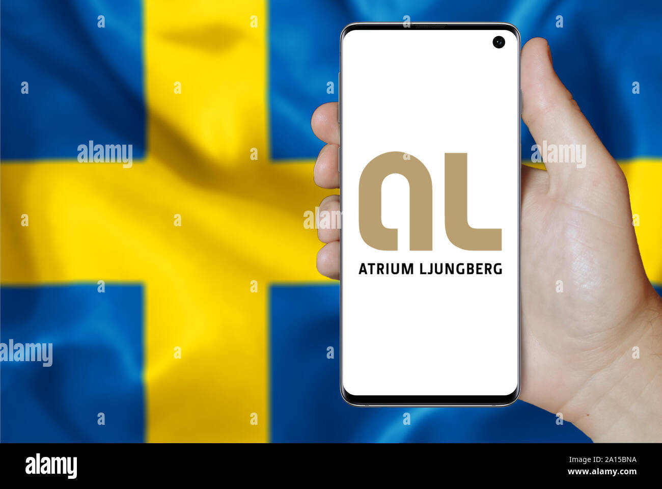 A man holds a smartphone displaying the logo of company Atrium Ljungberg listed on OMX Stockholm. Swedish flag in the background. Credit: PIXDUCE Stock Photo