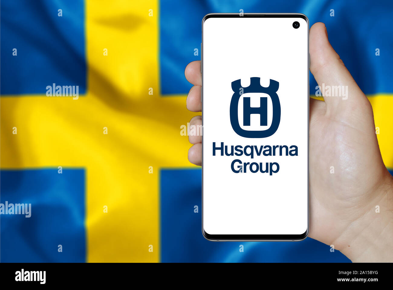 A man holds a smartphone displaying the logo of company Husqvarna listed on OMX Stockholm. Swedish flag in the background. Credit: PIXDUCE Stock Photo