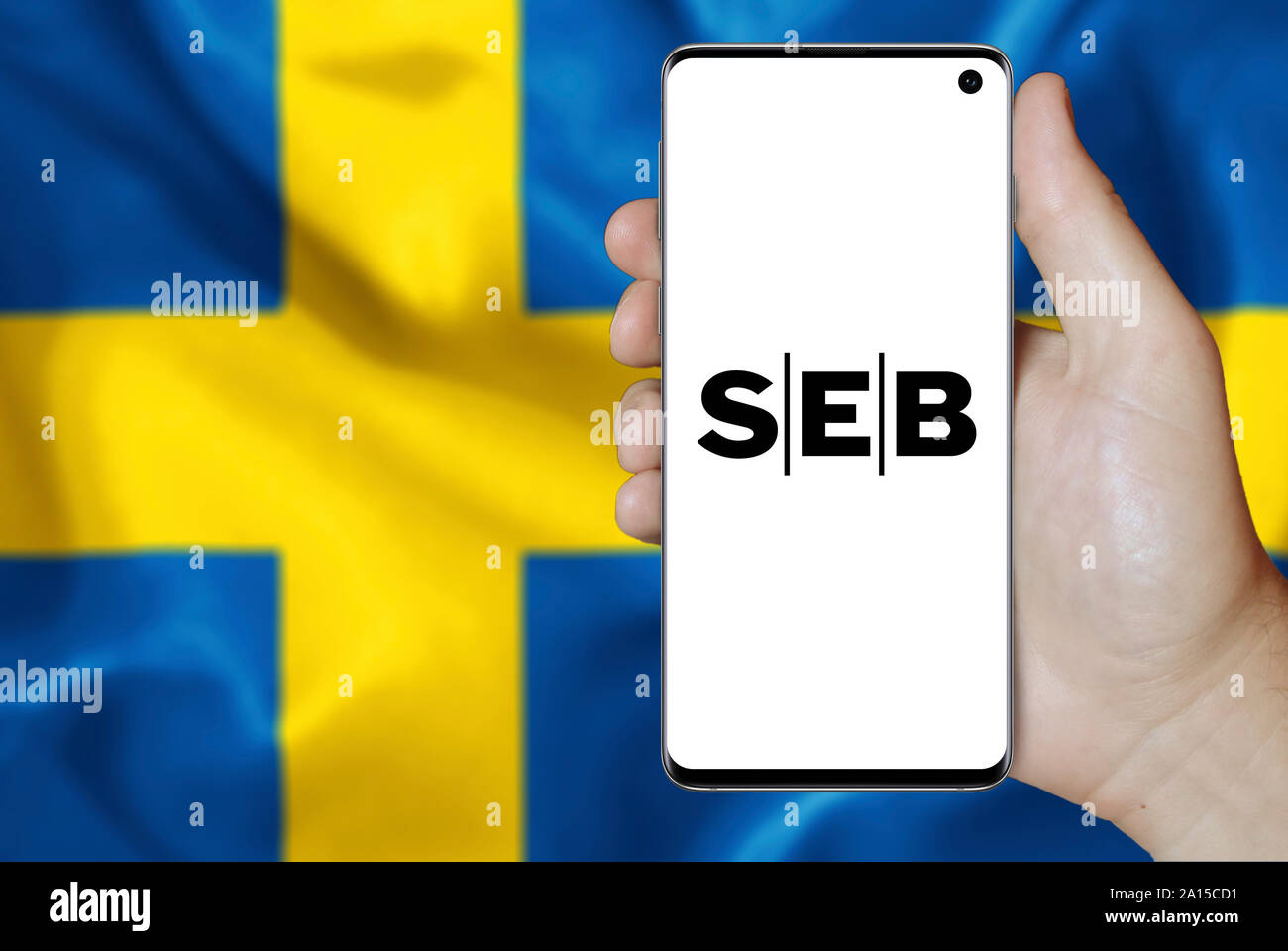 A man holds a smartphone displaying the logo of company SEB listed on OMX Stockholm. Swedish flag in the background. Credit: PIXDUCE Stock Photo