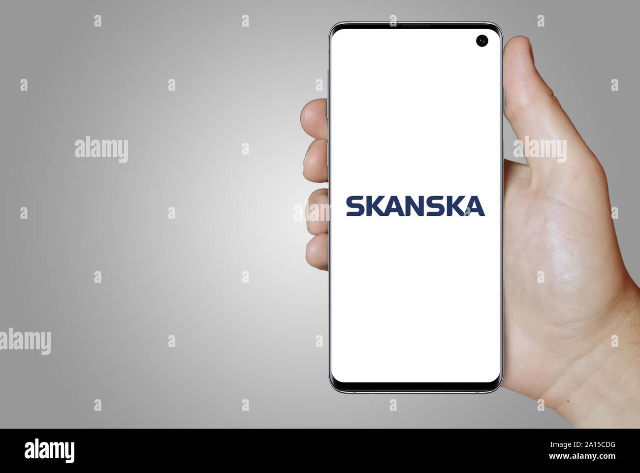 A man holds a smartphone displaying the logo of company Skanska listed on OMX Stockholm. Grey gradient in the background. Credit: PIXDUCE Stock Photo
