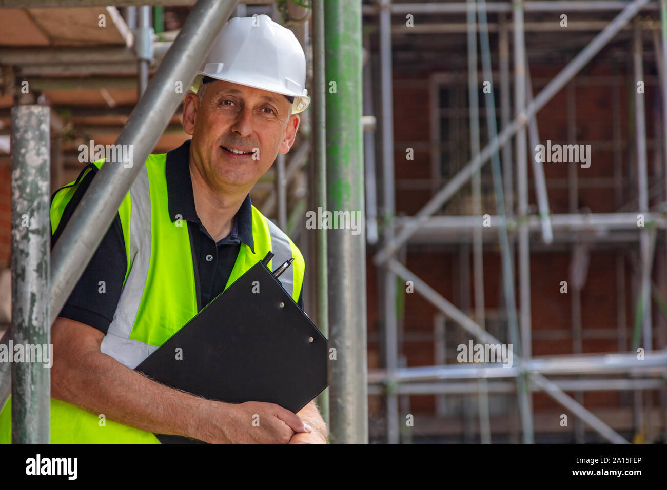 Male builder foreman, construction worker, engineer, surveyor or site manager holding a clipboard, wearing a white hard hat and hi vis vest standing o Stock Photo