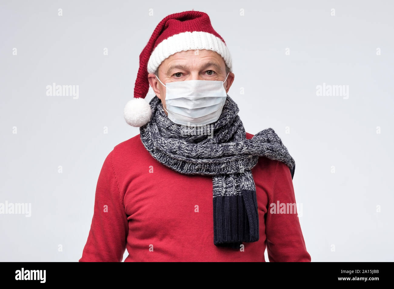 Caucasian mature man in red santa claus christmas hat closing face with medical mask protecting from virus. Studio shot Stock Photo