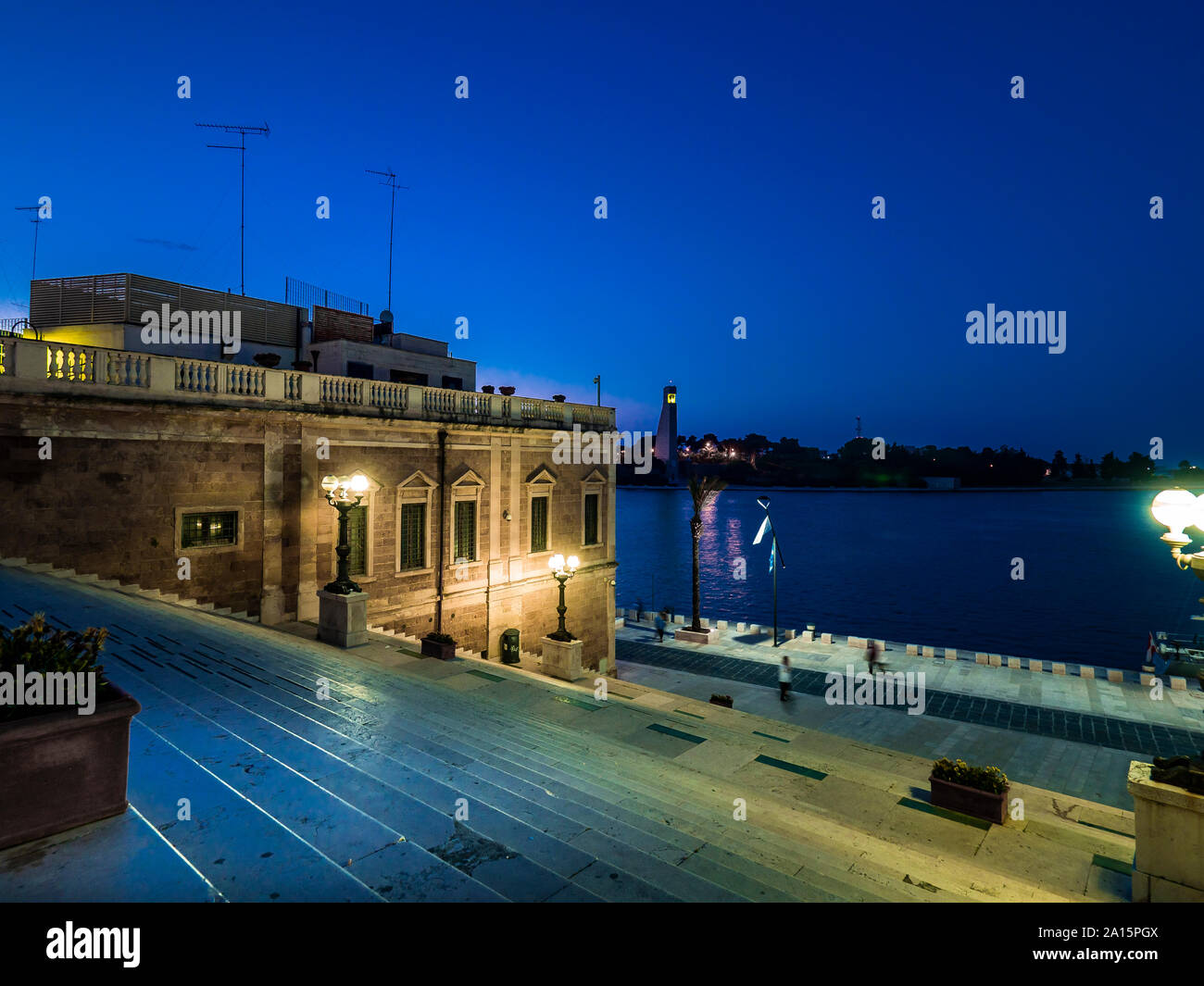 Illuminated Roman column on steps amidst buildings by sea in Brindisi against sky at night Stock Photo