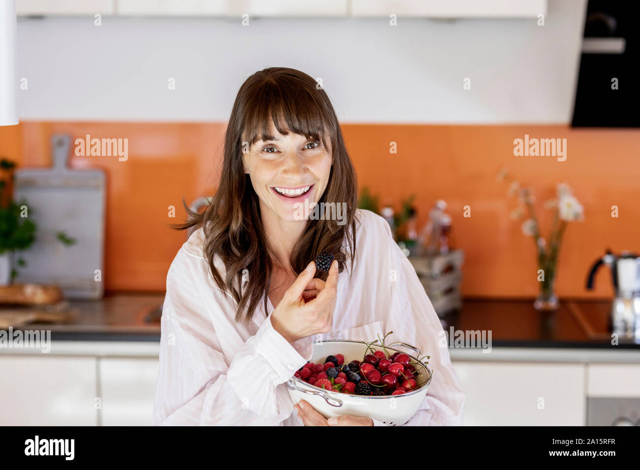 Portrait of happy woman wearing pyjama eating fruit in kitchen at home Stock Photo