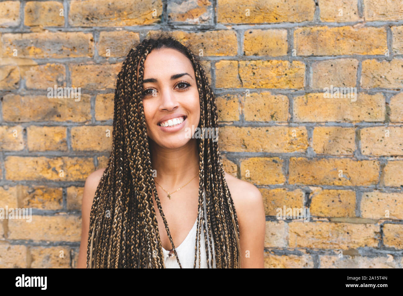 Portrait of happy young woman with long braids in front of brick wall Stock Photo