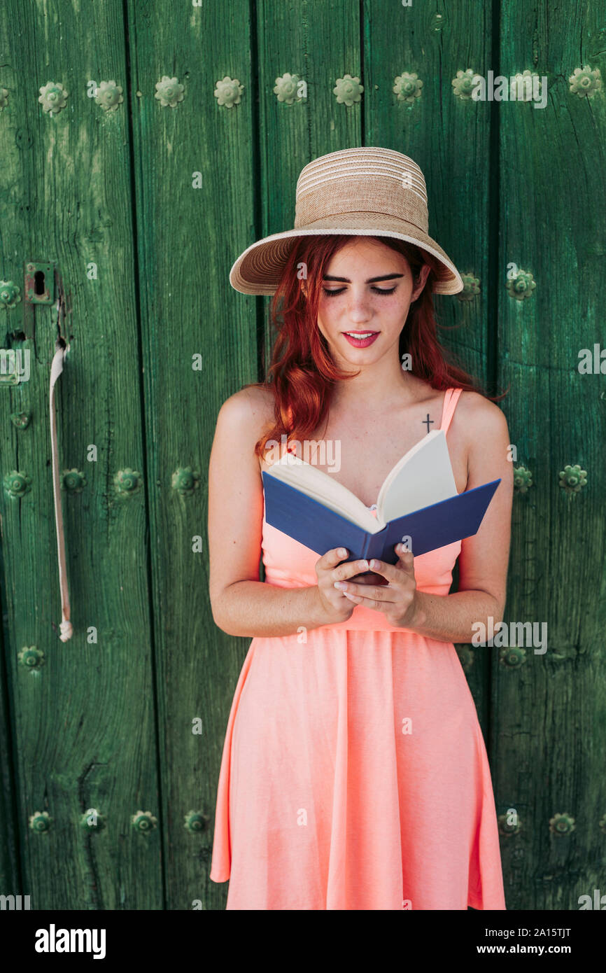 Portrait of redheaded young woman standing in front of green wooden door reading a book in summer Stock Photo