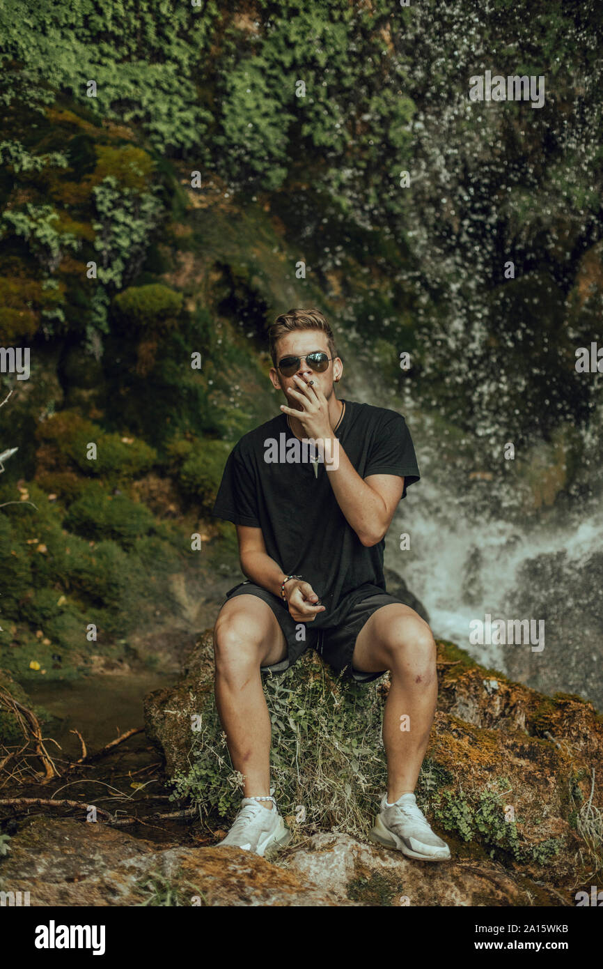 Portrait of a cool young man at a waterfall smoking a joint of marijuana Stock Photo