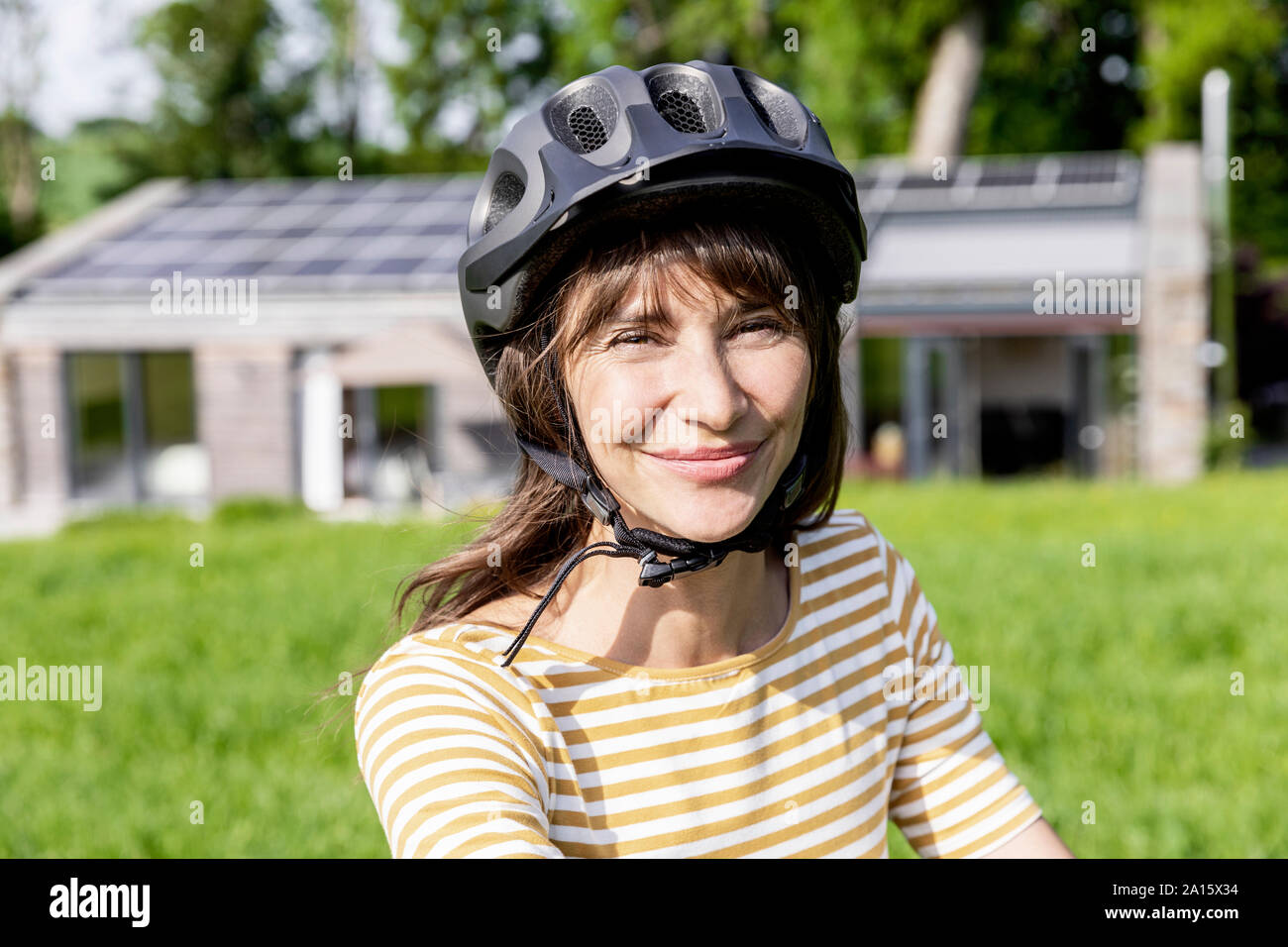Portrait of smiling woman with bicycle helmet on a meadow in front of a house Stock Photo
