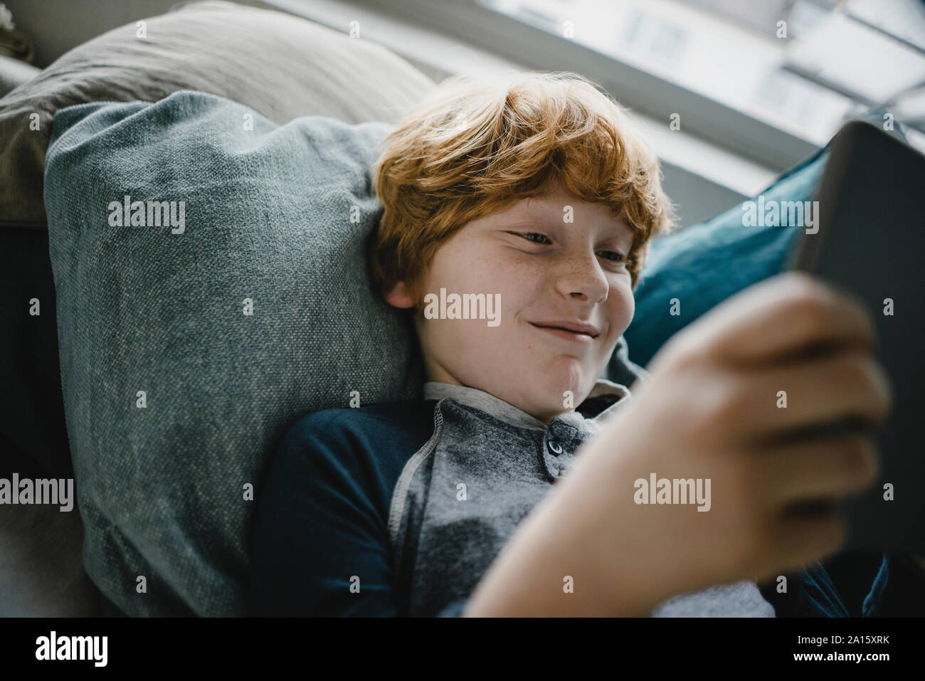 Portrait of smiling redheaded boy lying on couch using digital tablet Stock Photo
