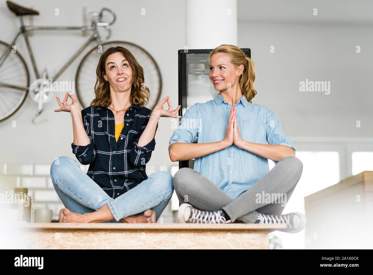 Two young women sitting cross-legged on the counter of their coffee shop, one meditating , other pulling funny faces Stock Photo