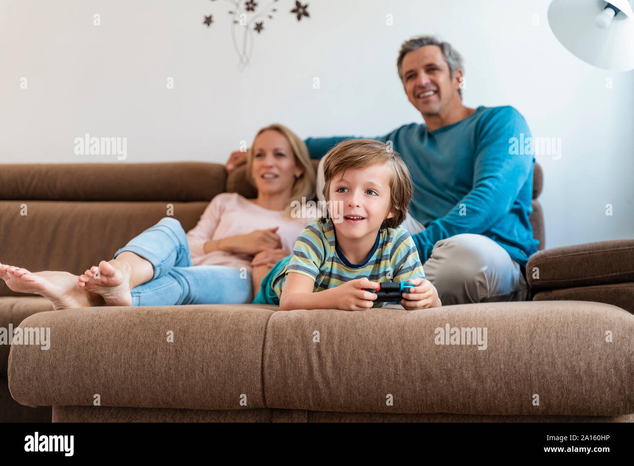 Boy lying on couch at home playing video game with parents watching Stock Photo