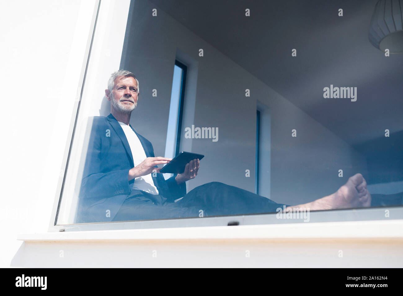 Senior businessman sitting at panorama window using tablet Stock Photo