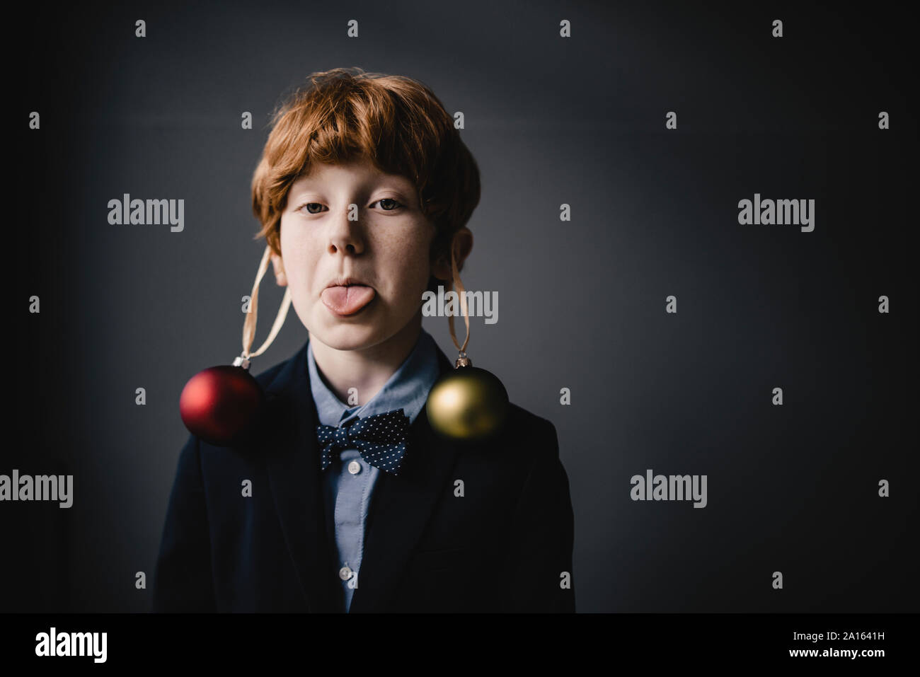 Portrait of redheaded boy wearing bow tie and Christmas baubles sticking out tongue Stock Photo