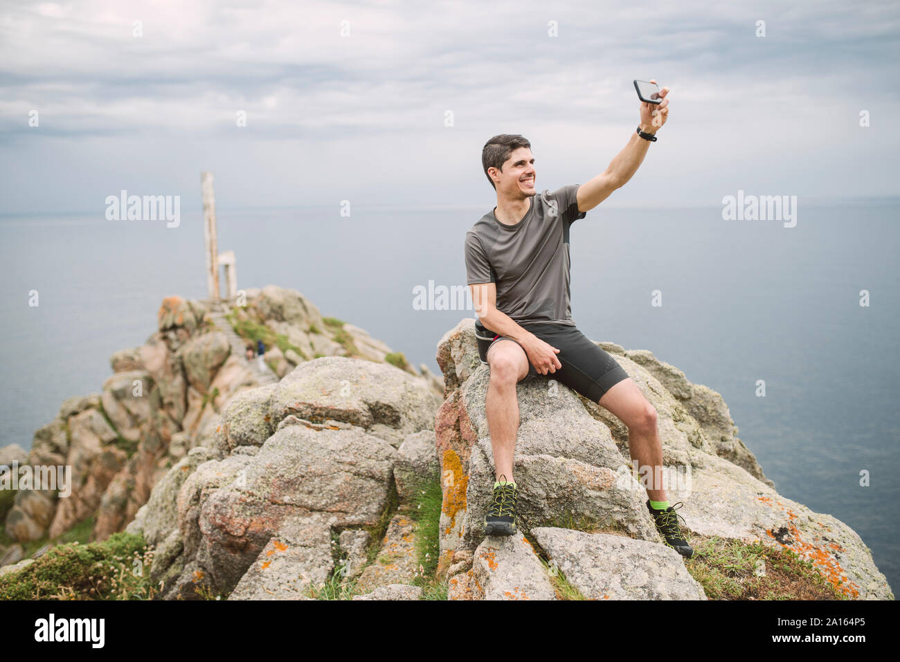 Trail runner sitting on a rock in coastal landscape taking a selfie, Ferrol, Spain Stock Photo