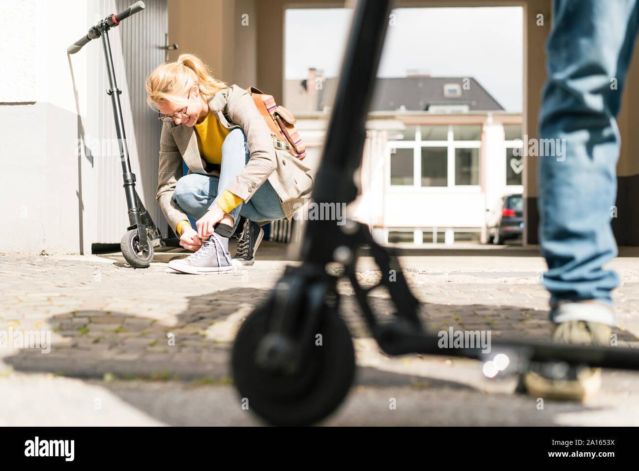 Young woman tying shoes, while friend is waiting with e-scooter Stock Photo