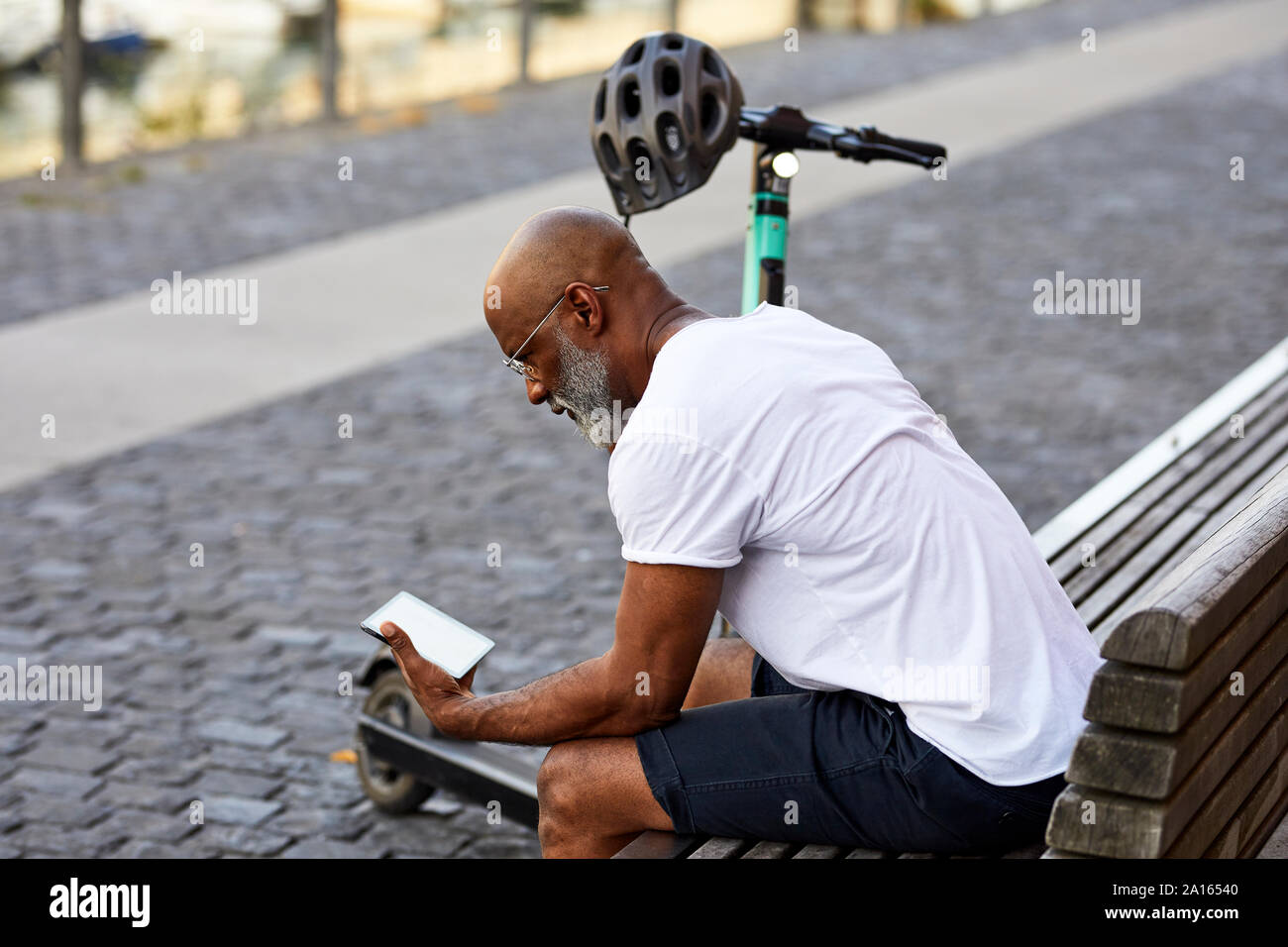 Mature man with E-Scooter sitting on bench in summer using mini tablet, Cologne, Germany Stock Photo