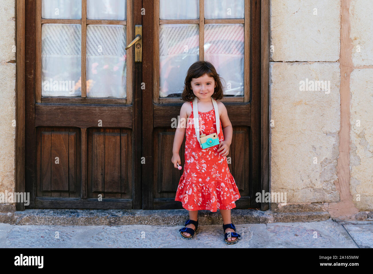 Portrait of smiling little girl with wooden toy camera wearing red dress with floral design Stock Photo