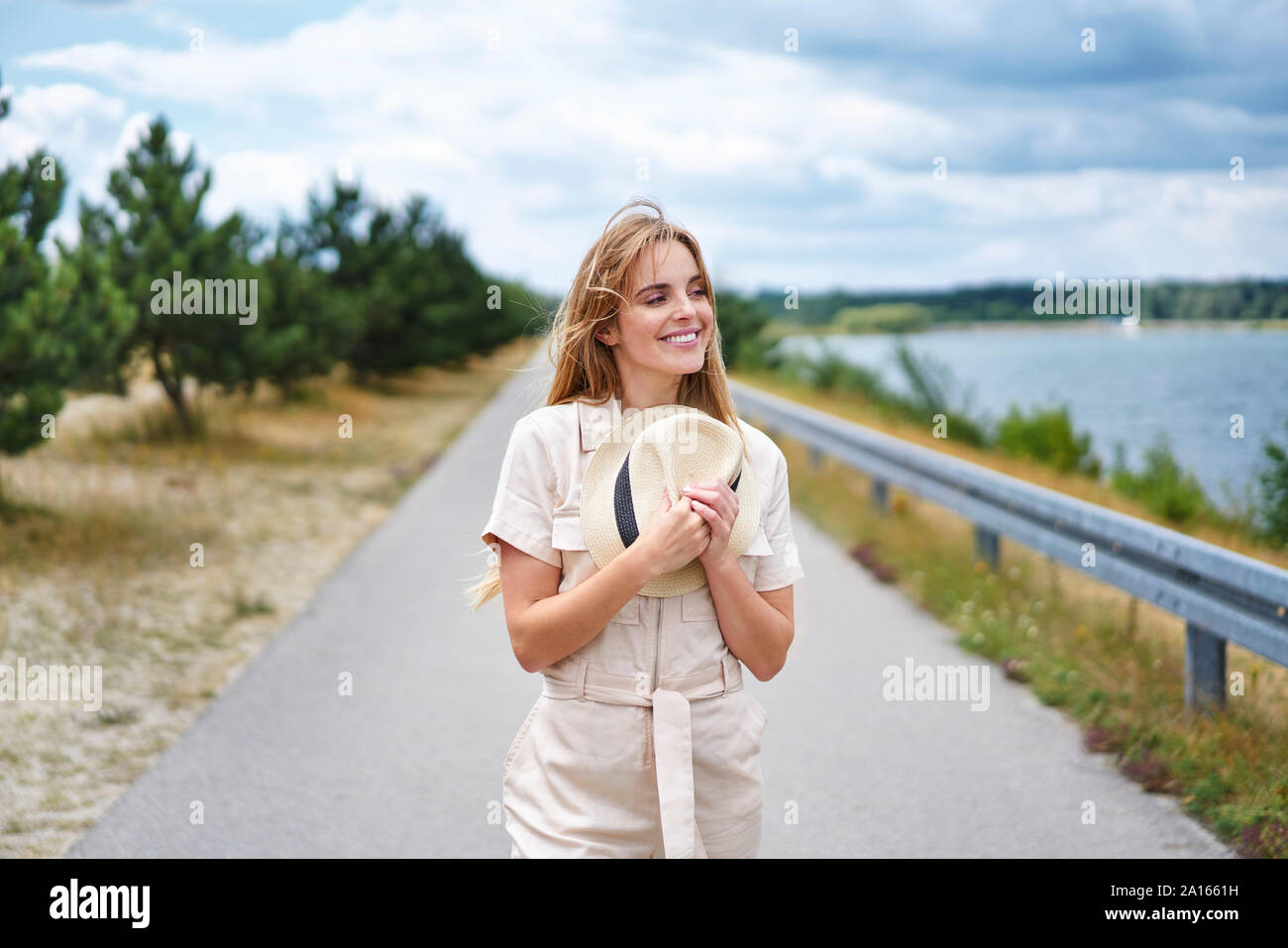 Smiling woman holding hat on rural road at the lakeside Stock Photo