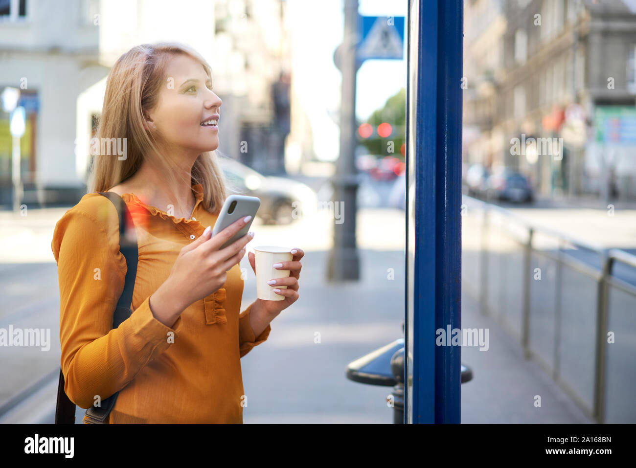 Smiling young woman with smartphone and takeaway coffee checking the schedule at bus stop Stock Photo