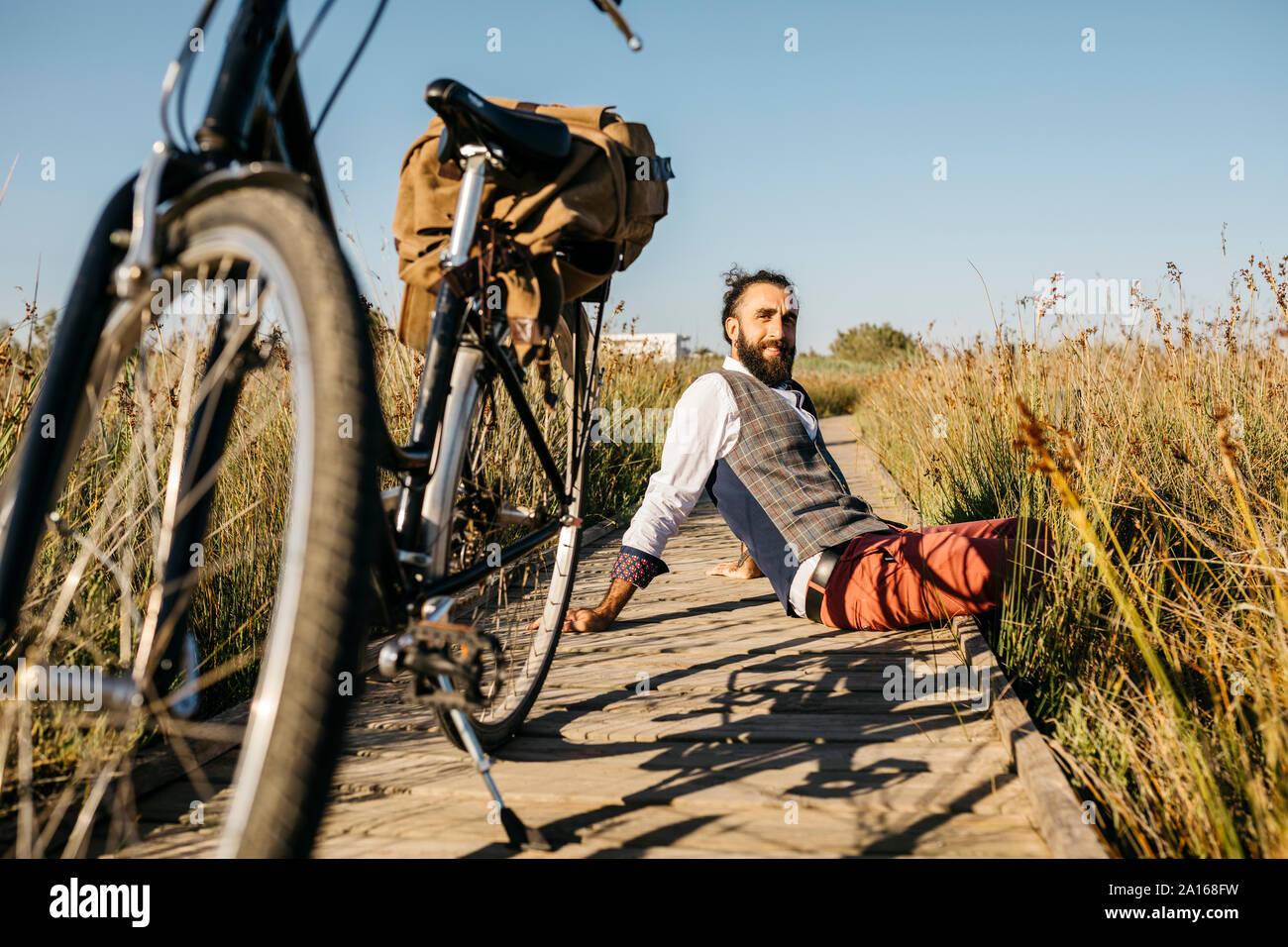 Well dressed man sitting on a wooden walkway in the countryside next to a bike Stock Photo