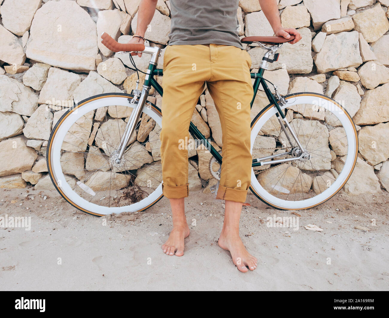 Barefoot man with Fixie bike in front of natural stone wall on the beach, partial view Stock Photo