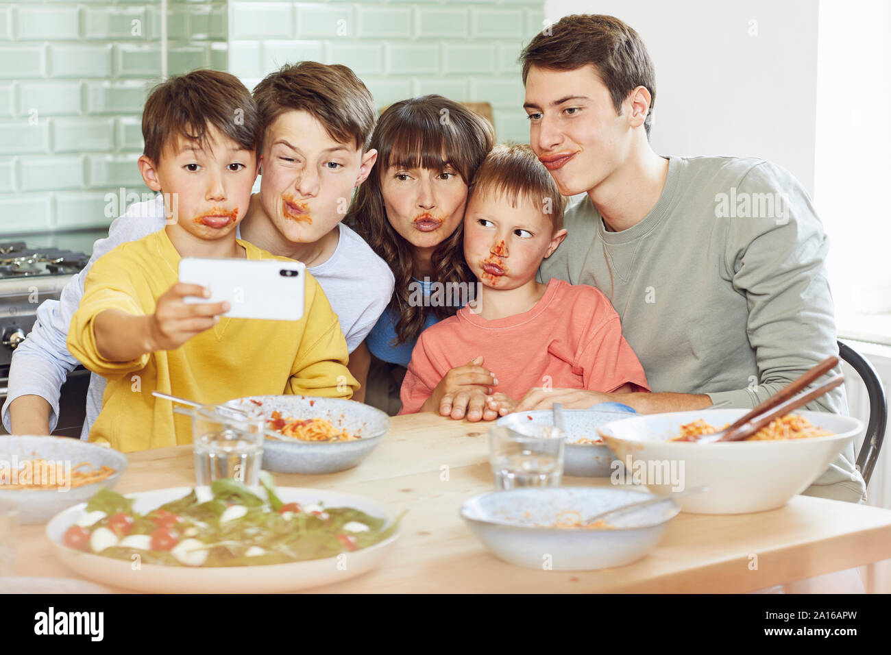 Mother and her four sons taking smartphone selfies at lunch, with faces full of tomato sauce Stock Photo