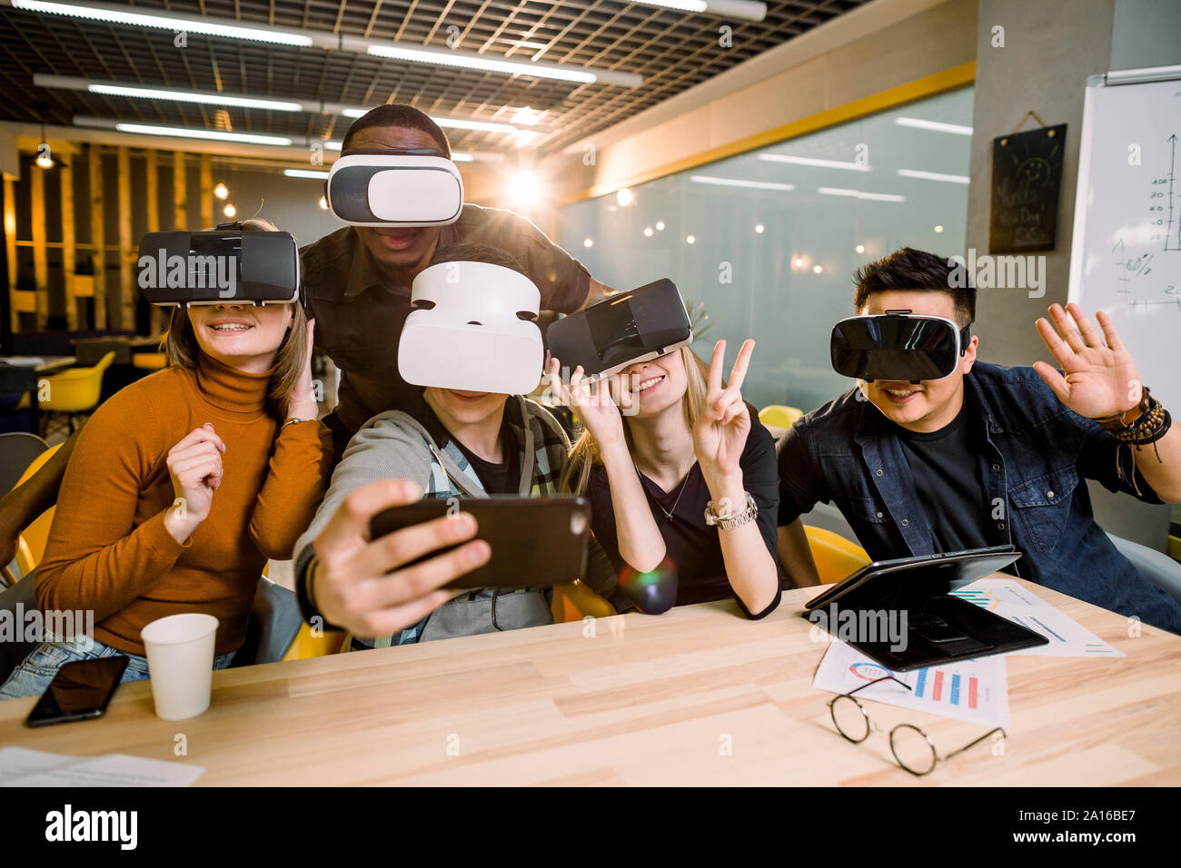 Five young funny people sitting at the table in front of each other, using virtual reality goggles, having fun and gesturing. VR goggles concept Stock Photo