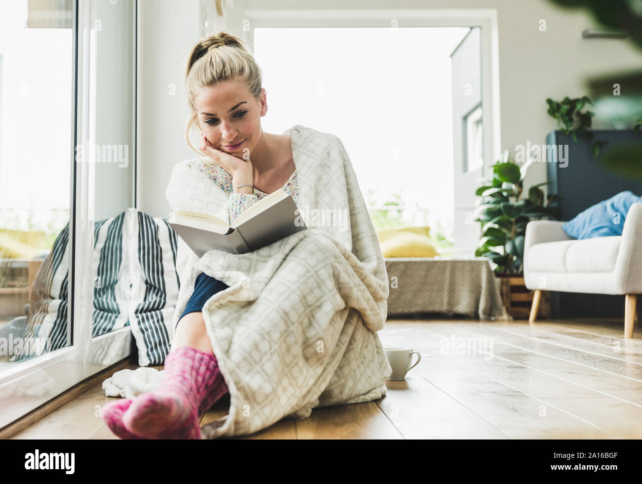 Woman wrapped in a blanket sitting at the window at home reading a book