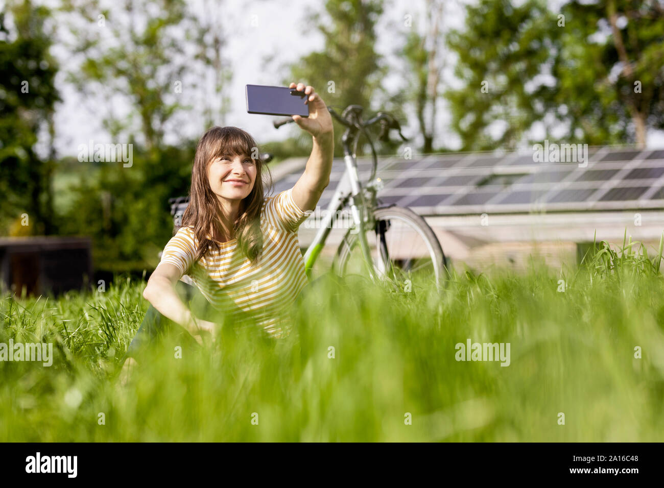 Relaxed woman sitting on a meadow with bicycle taking a selfie Stock Photo