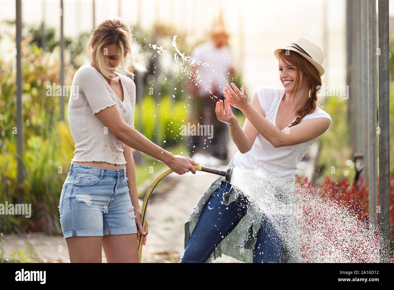 Two happy young women having fun while watering flowers with hose in the greenhouse Stock Photo