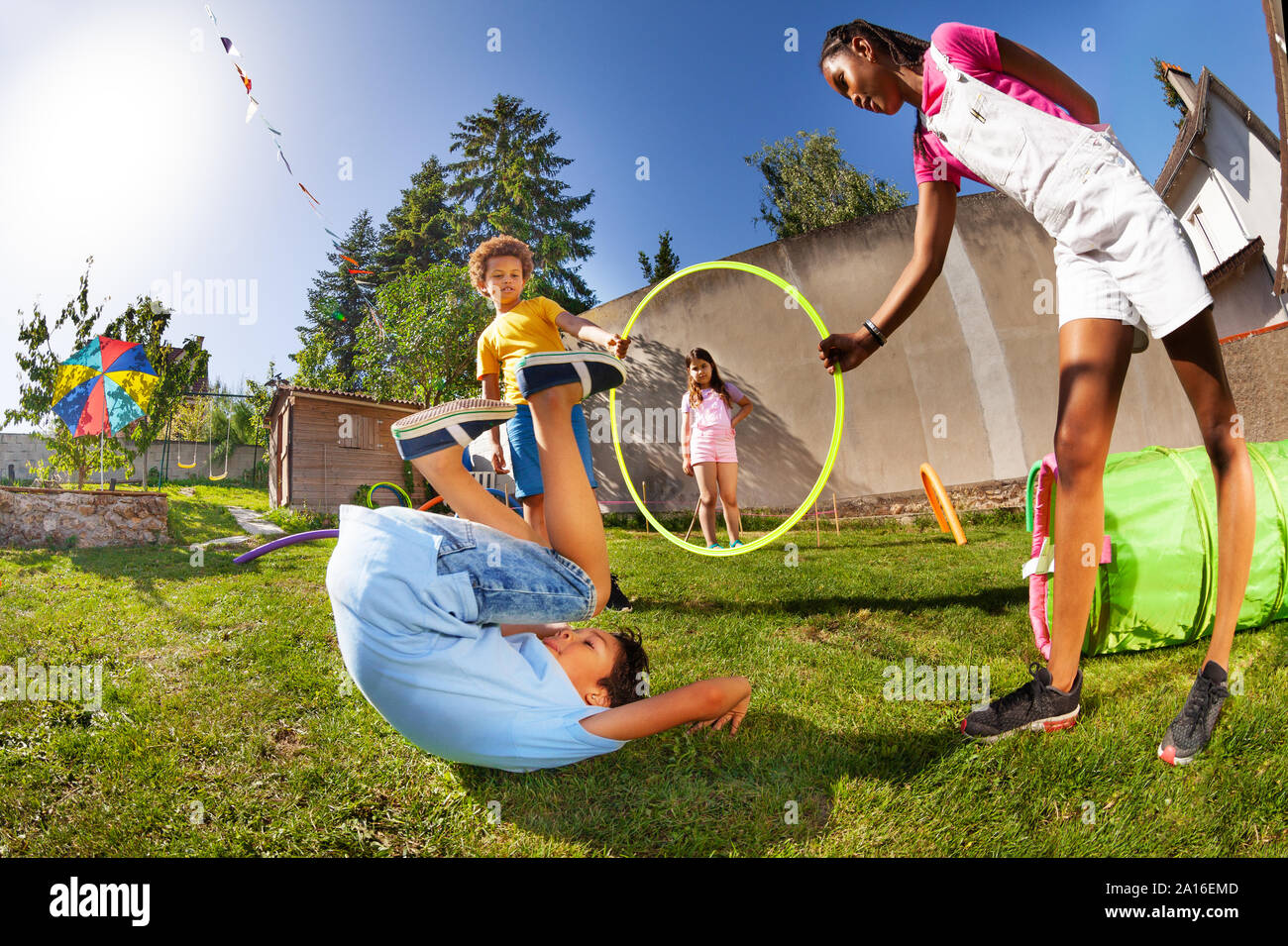 Happy boy jump through hula hoop ring hold by his friends on a playground lawn rolling on the grass after landing Stock Photo