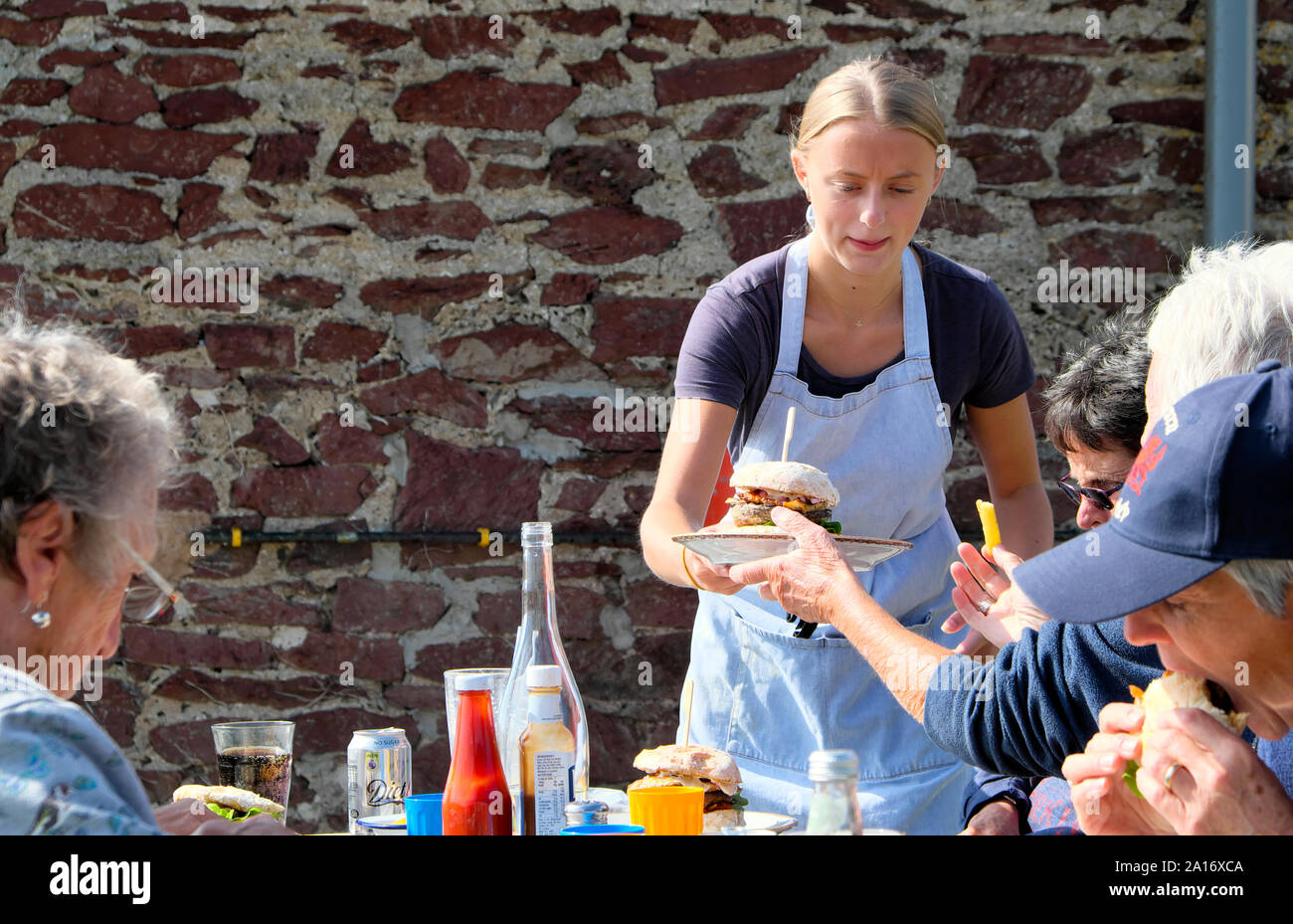 Young woman waitress serving burgers to customers eating outside at tables at Runwayskiln restaurant in Marloes Pembrokeshire Wales UK  KATHY DEWITT Stock Photo
