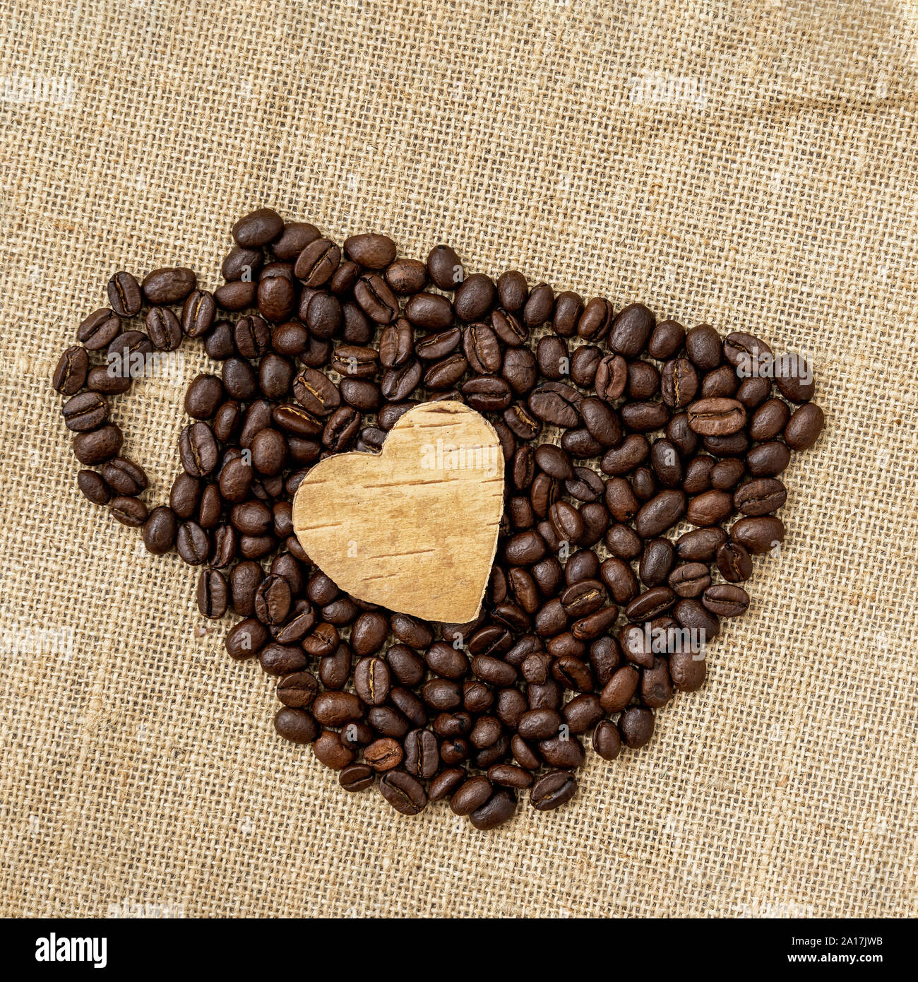 Coffee beans in the shape of a coffee cup with carved wooden heart on natural canvas, sackcloth. Concept of coffee love, loved one Stock Photo