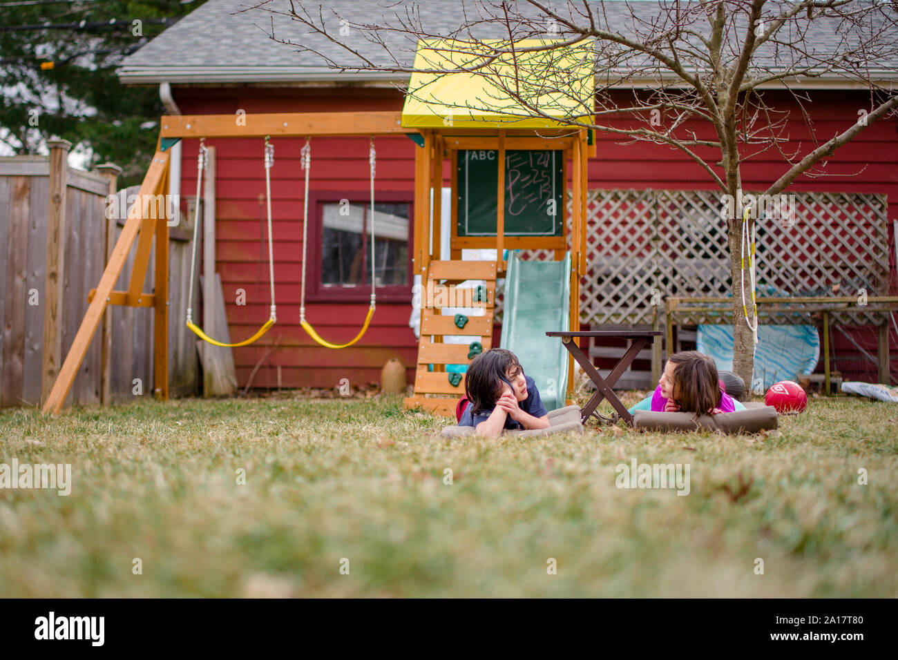 Two children lay in  grass in yard talking with playset in background Stock Photo
