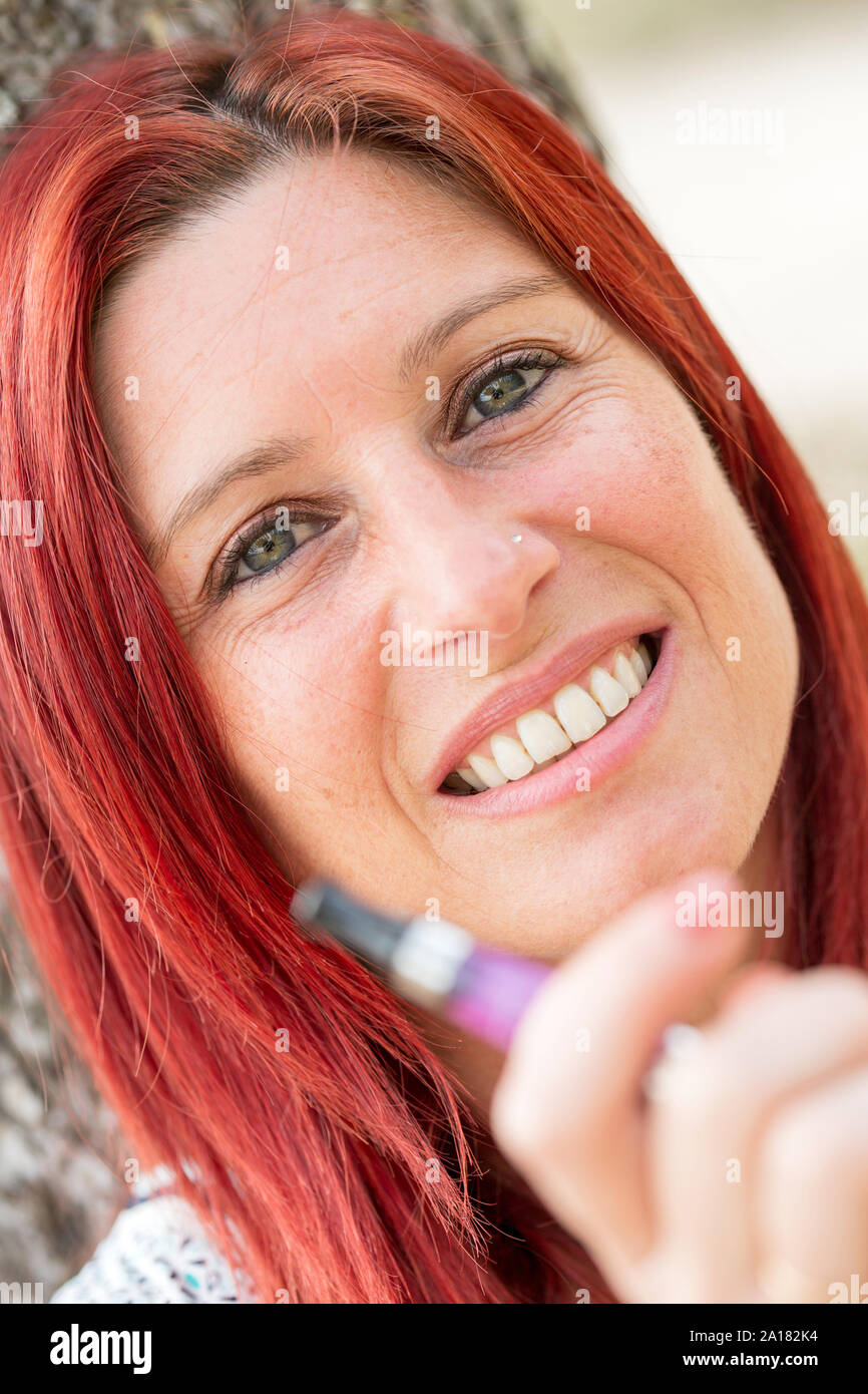 Portrait of a pretty redhead woman leaning against a tree smoking a electronic cigarette in a park Stock Photo