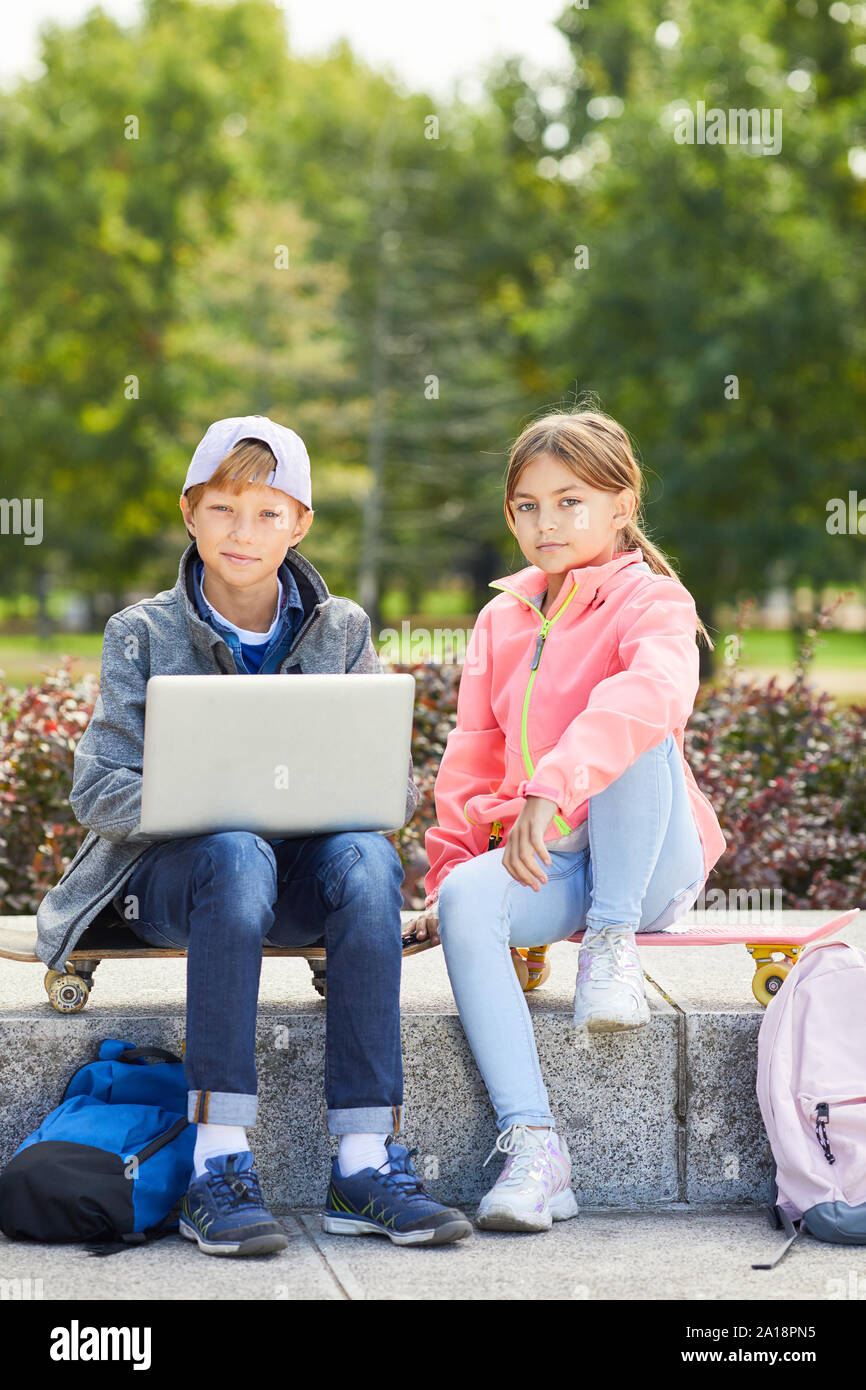 Portrait of boy and girl sitting on skateboards and looking at camera while using laptop computer outdoors in the park Stock Photo