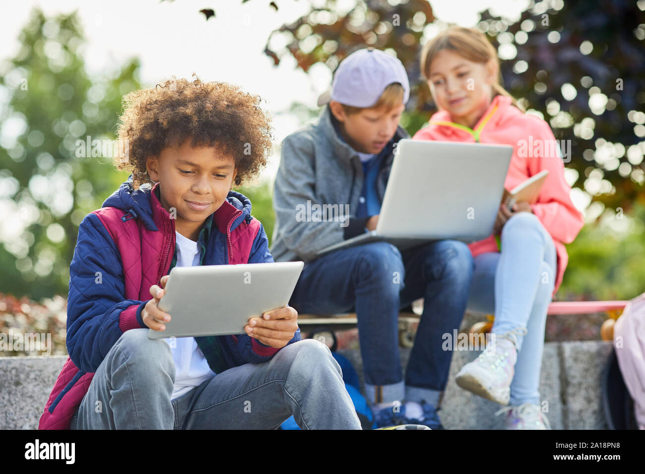African boy with curly hair sitting outdoors and using tablet pc with other children using laptop in the background Stock Photo