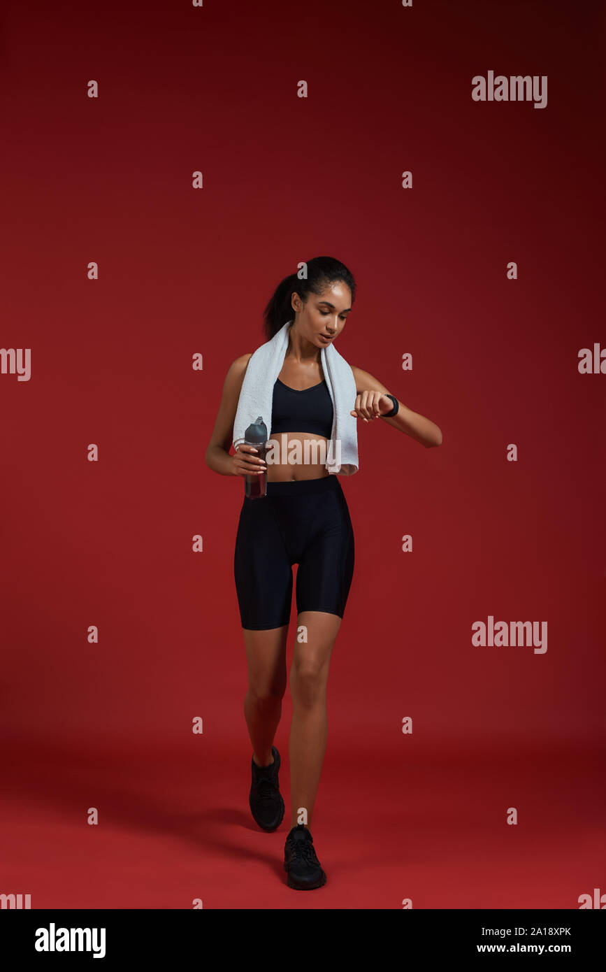 Great results. Full length of confident afro american women in sports clothing with towel on her shoulders holding bottle of water and checking the time while standing against red background. Sport concept. Active lifestyle Stock Photo