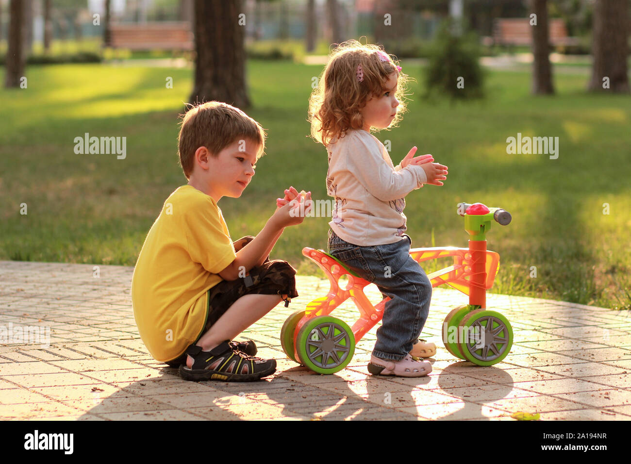 Little girl riding a small bike in the sunlit summer park and playing with a brother Stock Photo