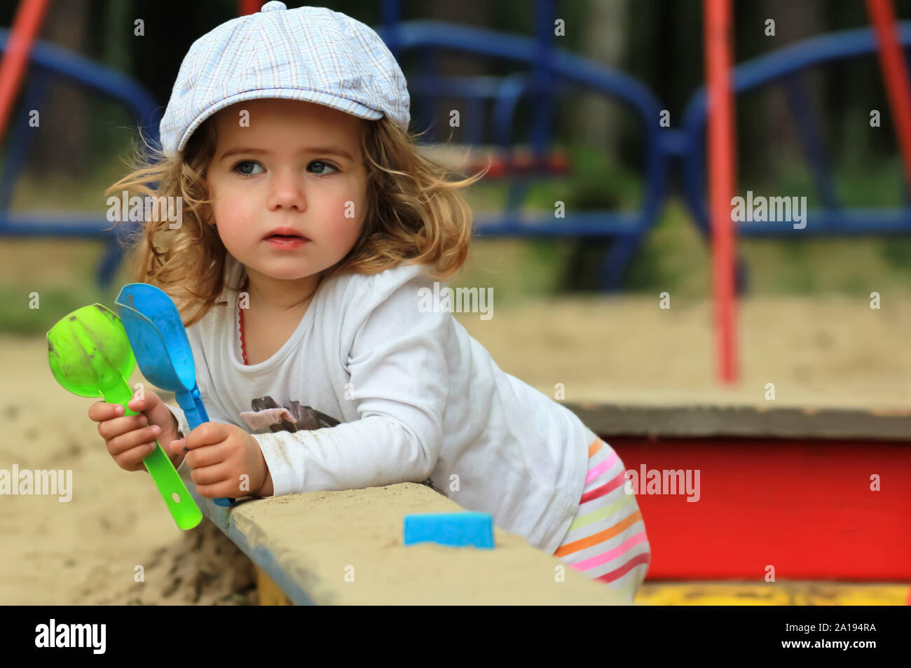 Thoughtful stylish one year old girl in a checked cap climbing out of a sandbox on a playground