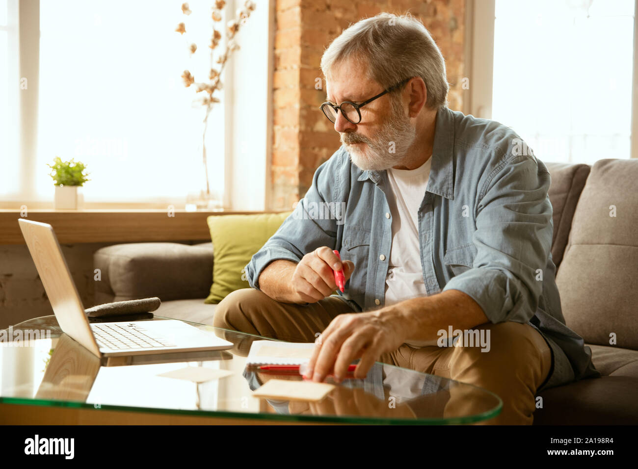 Senior man working with laptop at home - concept of home studying. Caucasian male model sitting on sofa and doing his homework or serfing in internet, watching cinema, making notes while webinar's on. Stock Photo