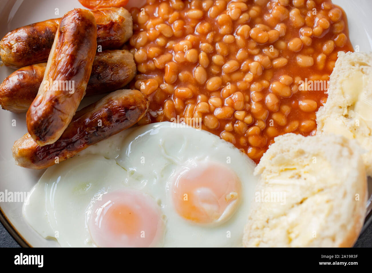 A meal of grilled pork sausages, grilled tomatoes, two fried eggs, baked beans and a buttered bread roll. Dark slate background. Dorset England UK GB Stock Photo