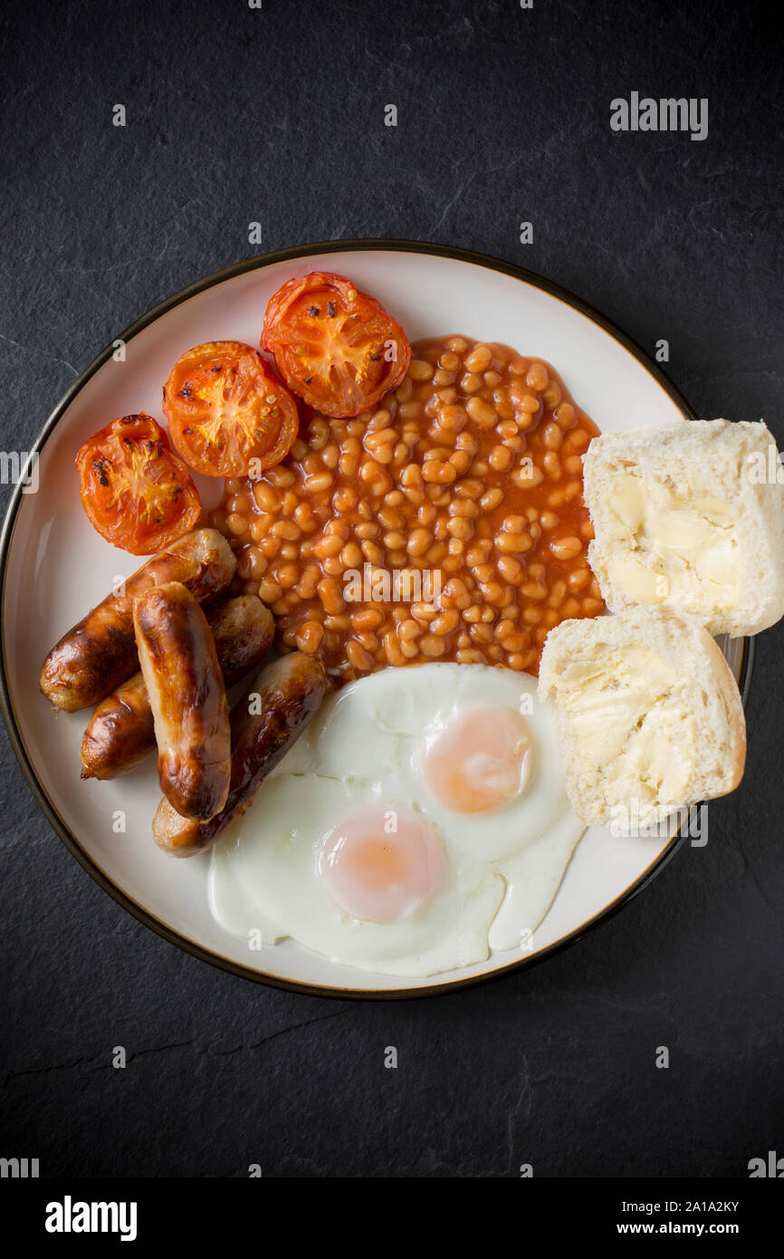 A meal of grilled pork sausages, grilled tomatoes, two fried eggs, baked beans and a buttered bread roll. Dark slate background. Dorset England UK GB Stock Photo