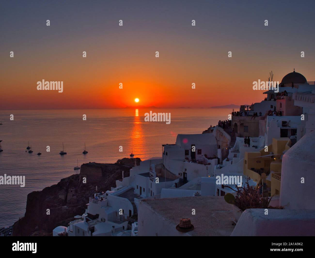 Majestic view of orange sun setting over tranquil water with boats and old town of Oia on Santorini island in Greece Stock Photo