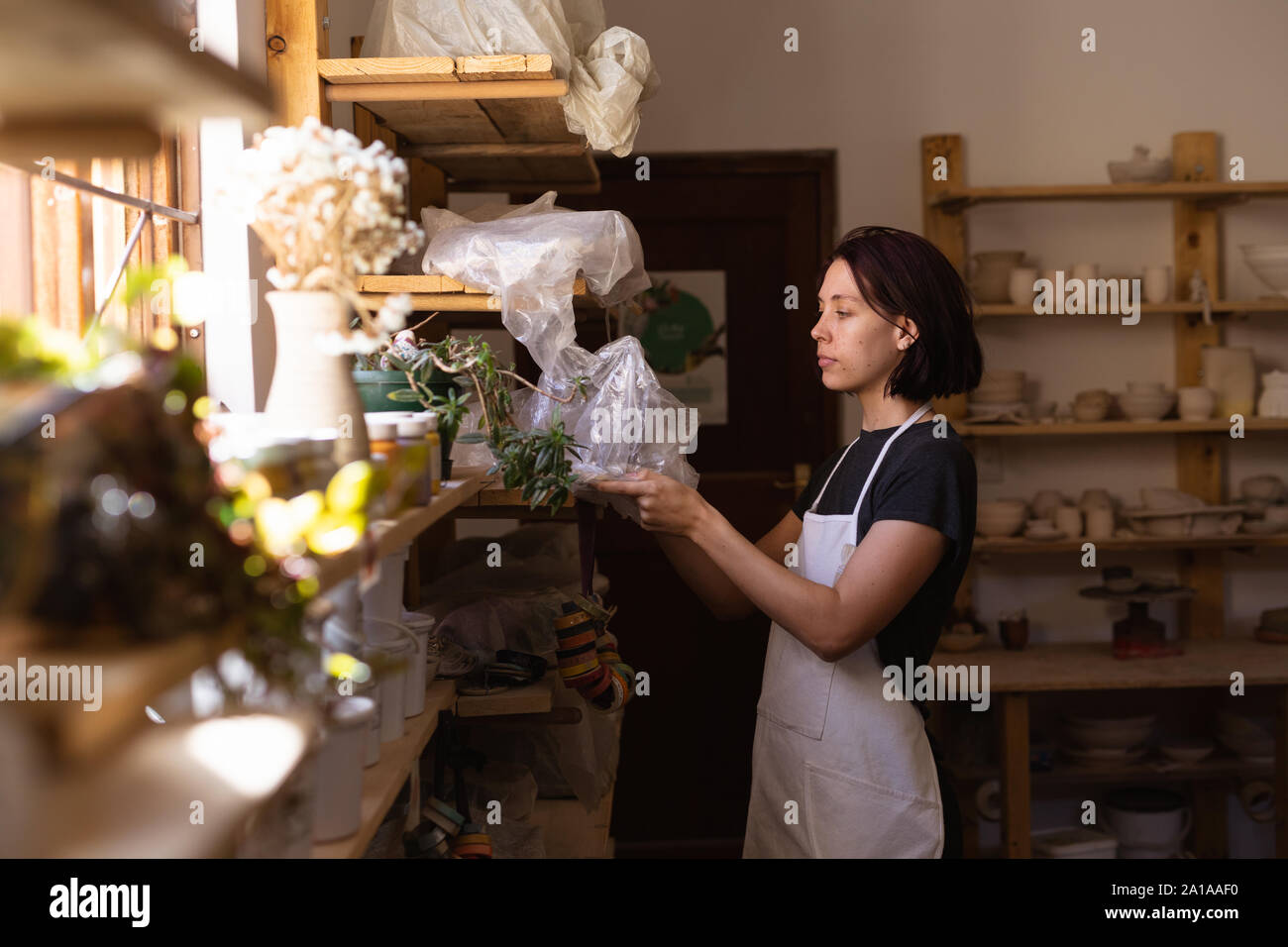 Female potter in a pottery studio taking down a bag of clay from a shelf Stock Photo