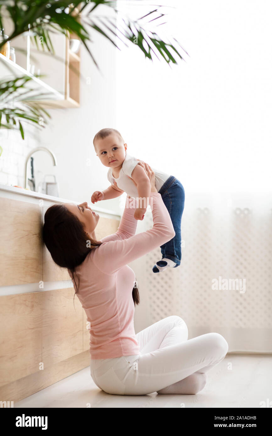 Young mom lifting her baby son up in air, sitting on kitchen floor