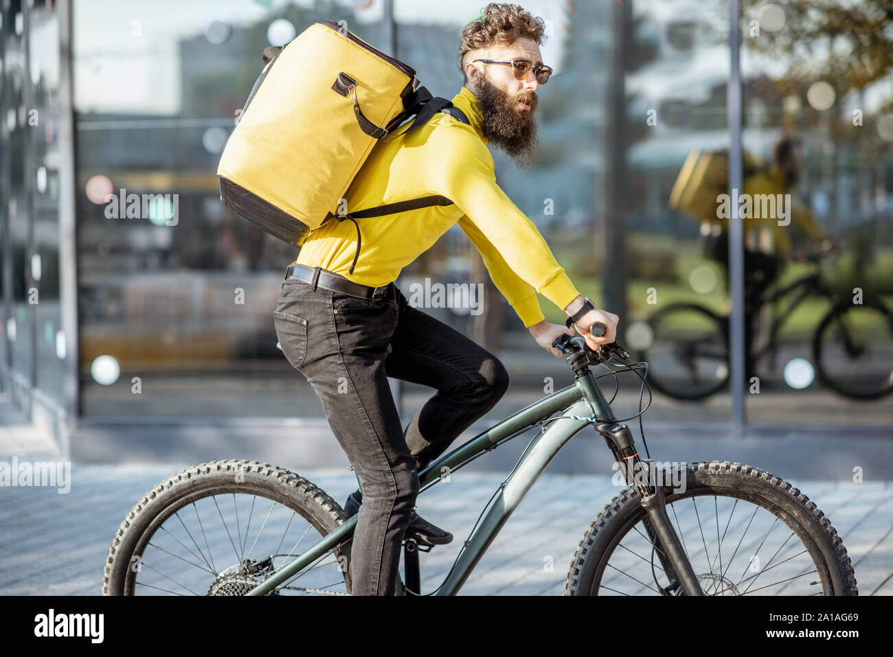 Young bearded courier delivering food with a yellow thermal backpack, riding a bicycle in the city. Food delivery service concept Stock Photo