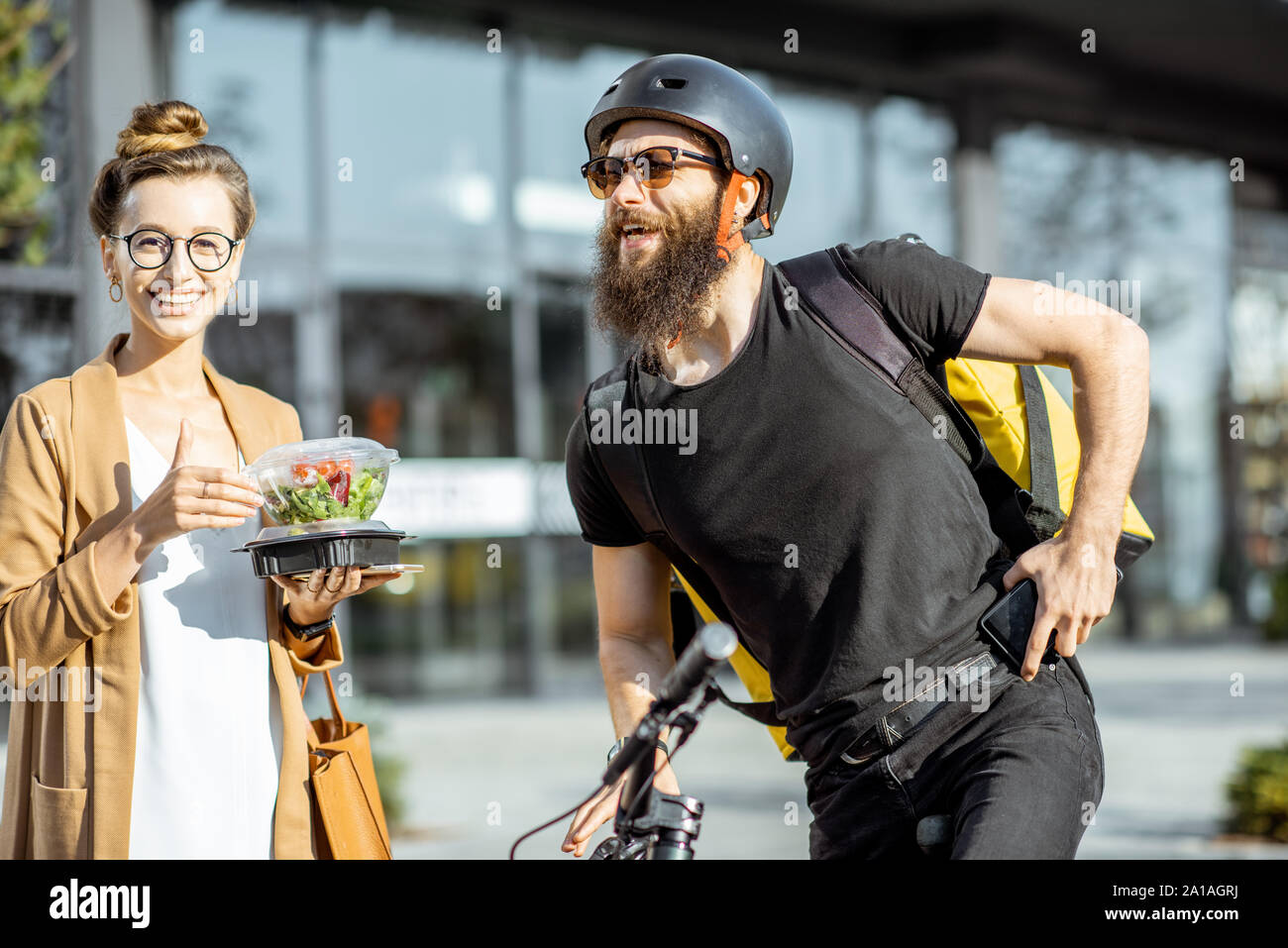 Happy businesswoman receiving fresh takaway lunches, standing near the office building with a male courier on the background. Takeaway restaurant food Stock Photo