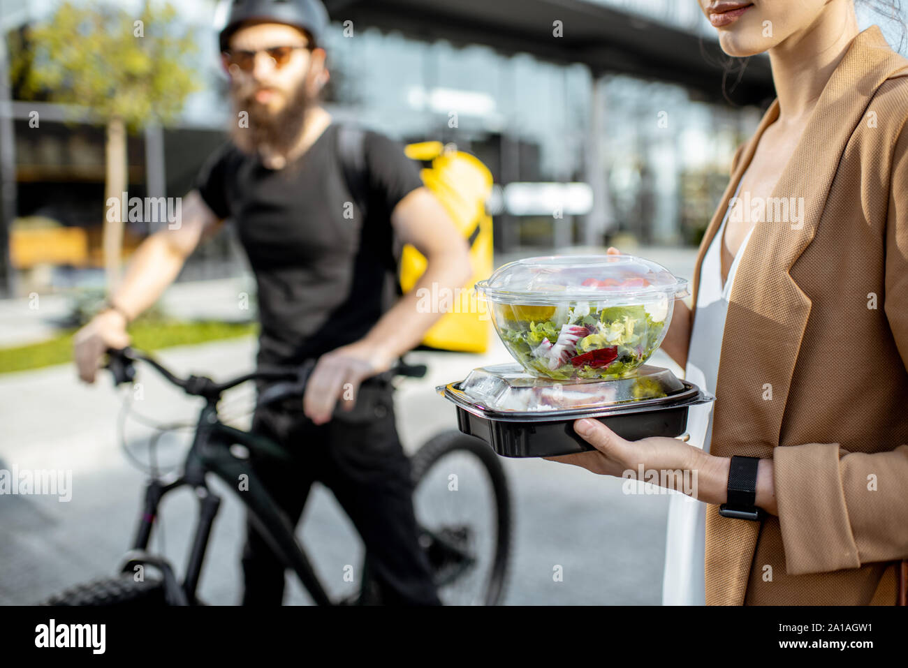Businesswoman holding lunchboxes with fresh takaway food outdoors. Male courier on a bicycle on the background. Takeaway restaurant food delivery conc Stock Photo